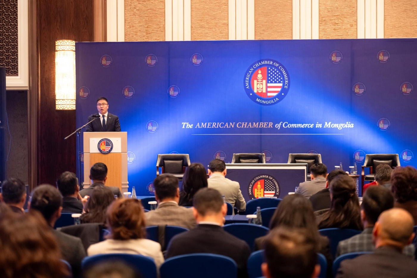 A speaker at a lectern in front of the AmCham Mongolia seal, with the audience seen from behind.